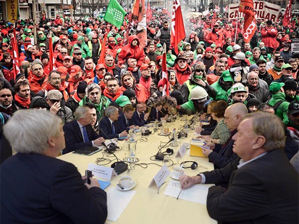 Fotomontage overlegtafel met bestuurders waar achter een demonstratie oprukt